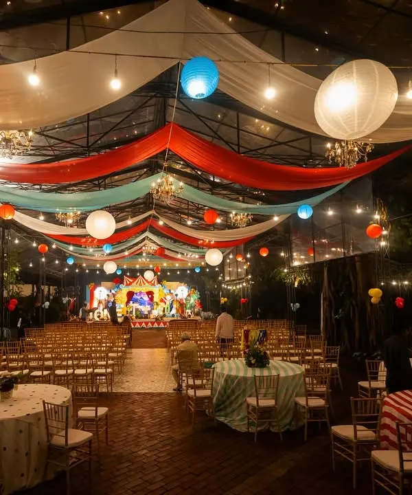 A wide shot of a spectacular carnival-themed event at night. The venue is transformed with colorful draped fabrics, paper lanterns, and string lights, creating the festive atmosphere of a big top tent.