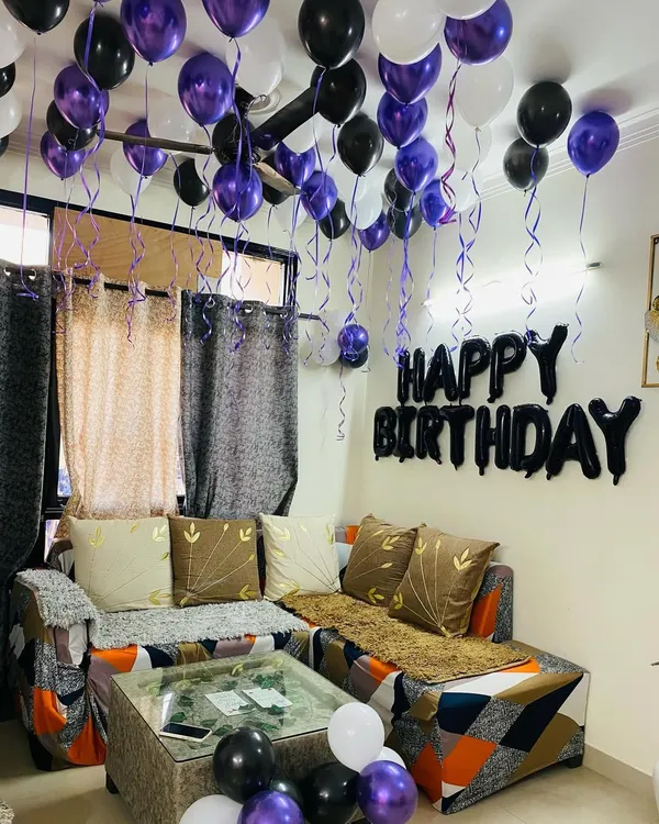 A simple and classic birthday decoration for a living room. This setup includes purple, black, and white balloons on the ceiling and a "Happy Birthday" foil banner on the wall.