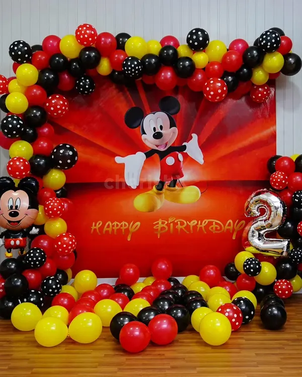 A classic Mickey Mouse theme for a 2nd birthday. This decoration features a large printed backdrop of Mickey, framed by a balloon garland in his signature red, yellow, and black colors, including some with polka dots. A large number '2' foil balloon and a Mickey Mouse balloon figure complete the look.
