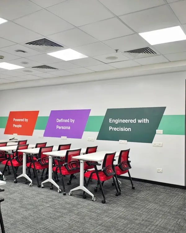 A flexible training room at the Mindsprint office with movable tables and stackable chairs. The wall graphics reinforce the company's core principles: "Powered by People, Defined by Persons, Engineered with Precision."
