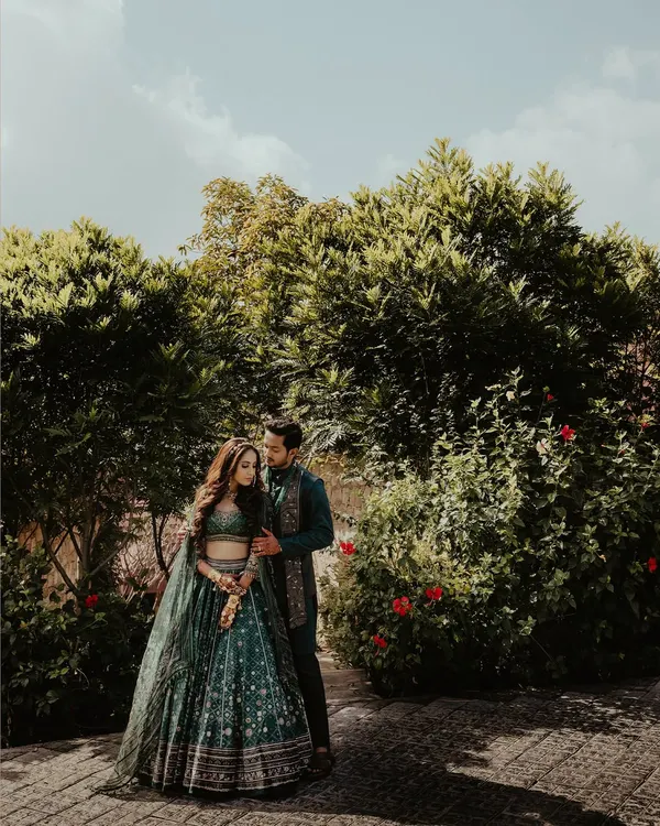 A beautiful wide shot of a couple in coordinated dark green traditional outfits, standing in a lush garden. The natural lighting and rich colors make this a stunning pre-wedding portrait.
