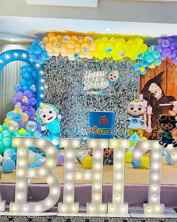 A vibrant Cocomelon-themed first birthday party. This stage setup features a silver shimmer wall, character cutouts, a tall giraffe prop, and large marquee letters.