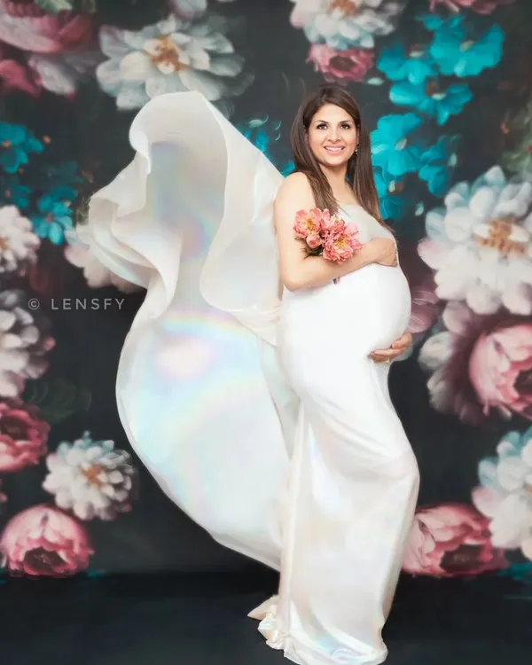 A fine art maternity portrait against a rich floral backdrop. The flowing white fabric of the gown adds a touch of drama and elegance, creating a timeless, painterly photograph to celebrate your pregnancy.