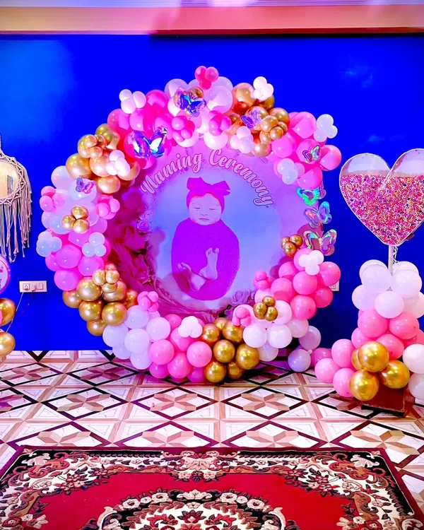 A stunning decoration for a Naming Ceremony. This setup features a personalized backdrop with the baby's photo, framed by a beautiful ring of pink, white, and gold balloons with butterfly accents.