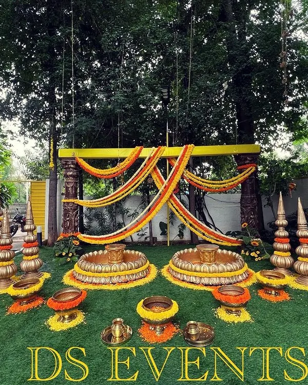 An elaborate outdoor setup for a Haldi or Pellikuthuru ceremony. This design features two large lotus-shaped urlis for seating, surrounded by traditional brass decor elements and marigold flower arrangements.