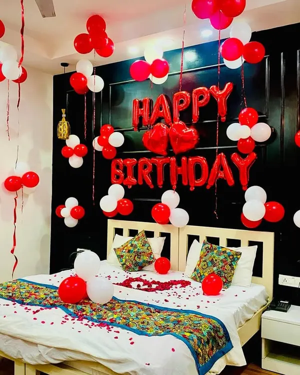 A classic romantic birthday bedroom decoration. The room is filled with red and white balloons, a large "Happy Birthday" foil balloon set above the bed, and rose petals scattered for that lovely touch.