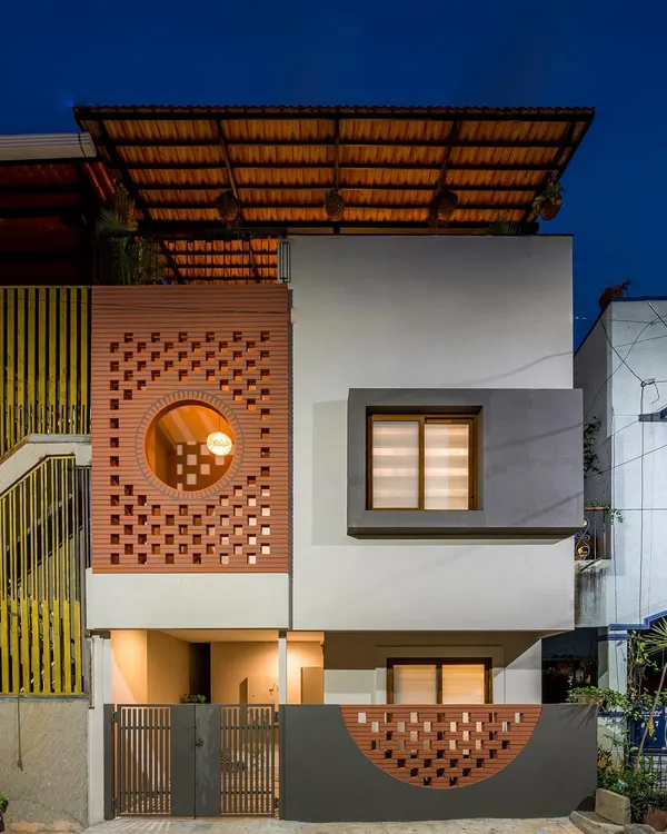 The facade of the Mathrukrupa residence at dusk. The perforated terracotta brick screen, a modern take on the traditional jali, is a key element for passive cooling, allowing air to flow while filtering harsh sunlight.