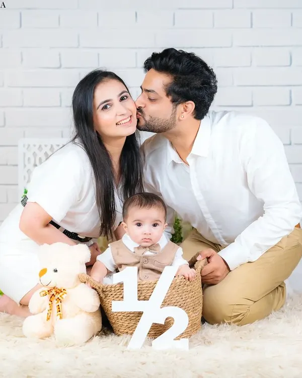 A sweet half-birthday celebration photo. The father's kiss on the mother's cheek while they surround their baby creates a moment of pure family love.