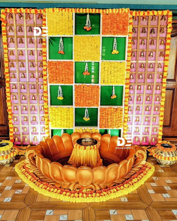 A traditional Haldi ceremony setup featuring a custom-made lotus-shaped seating area. The backdrop is a vibrant grid of green banana leaves and marigold flowers, accented with temple bells for an authentic touch.