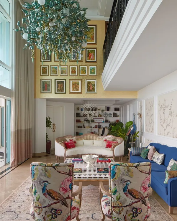 In this double-height South Bombay living room, we created multiple seating areas to make the large space feel intimate. A gallery wall draws the eye upward, while a mix of classic and modern furniture, like the floral armchairs and bold blue sofa, adds personality and color.