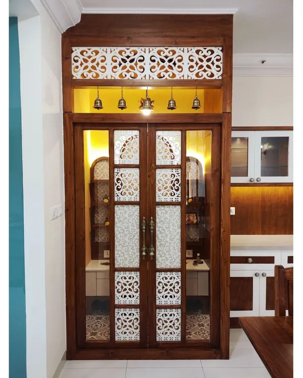 The entrance to the magnificent pooja room in #TheDivinityHome. The custom doors feature intricate jaali work, frosted glass panels, and traditional brass bells, creating a grand and welcoming threshold to this sacred space.
