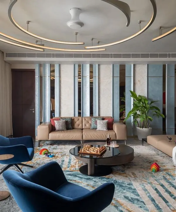 The living room of our Quintessence project in Bangalore, where design is inspired by the nearby Jakkur Lake. The custom ceiling lights mimic the movement of water ripples, while the open layout and large windows create a bright, expansive feel. This space combines luxurious leather sofas, vibrant blue armchairs, and a mix of textures to create a sophisticated yet comfortable family area.