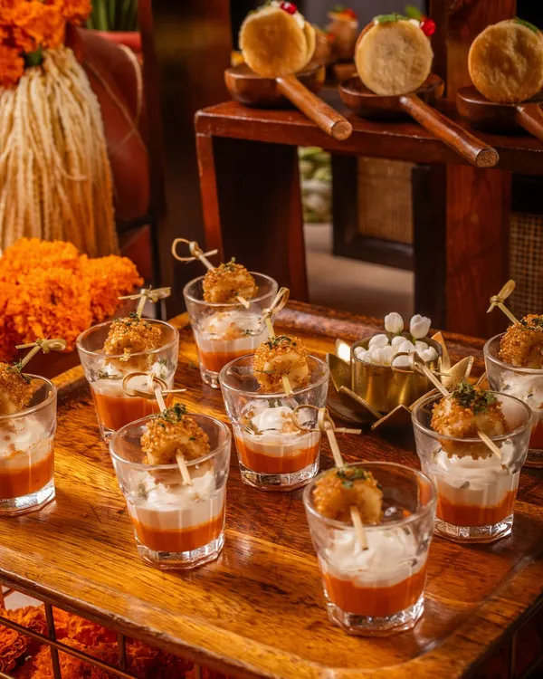 An array of our signature appetizers, perfect for a festive celebration. This tray features crispy bites in shot glasses with a layered dip and pani puris on custom wooden spoons, all set against a backdrop of vibrant marigold flowers.