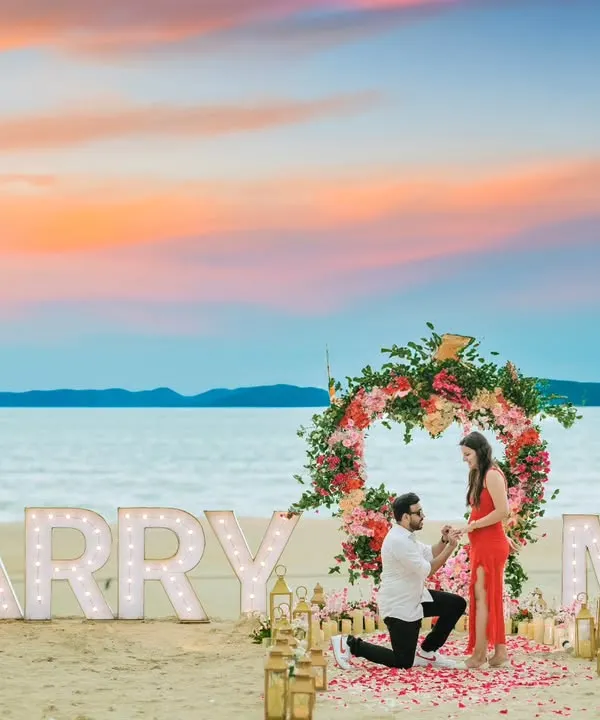 A picture-perfect beach proposal captured during a pre-wedding shoot. The "MARRY ME" marquee lights, floral arch, and stunning sunset sky create an unforgettable romantic moment.