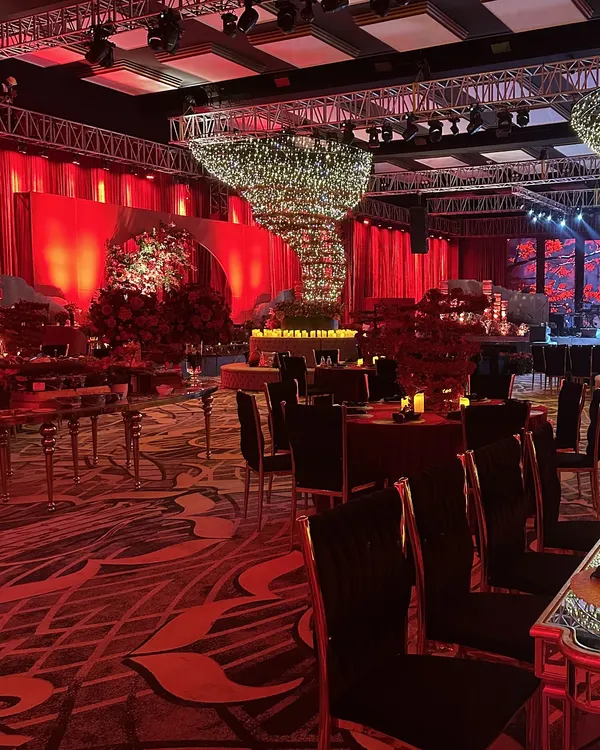 A wide shot of the sangeet at Fairmont Mumbai, bathed in a dramatic red glow. You can see the scale of the event, from the velvet-draped walls to the statement flower tornado chandelier, all working together to create an immersive world of color.