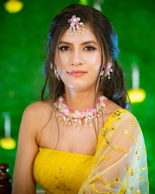 The Haldi glow is real. This is a beautiful close-up portrait of a bride-to-be, adorned with delicate floral jewelry, looking directly at the camera with a serene smile. The vibrant green background makes her yellow outfit pop.