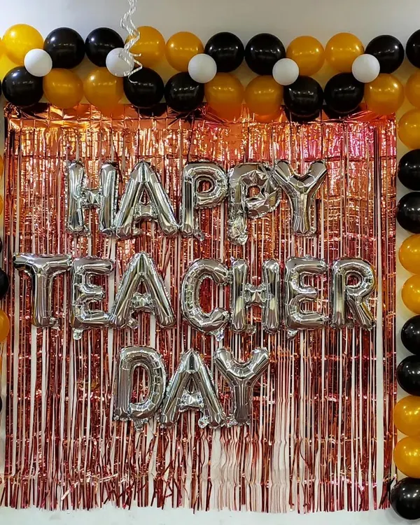 A classic backdrop for an office Teacher's Day celebration. This setup features a rose gold tinsel curtain, 'Happy Teacher Day' silver foil balloons, and a neat frame of black, gold, and white balloons, creating a perfect spot for photos.
