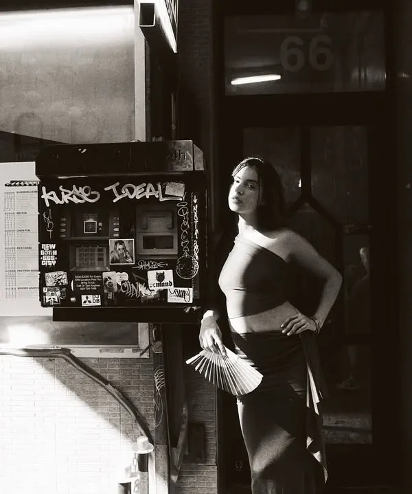 A black and white street portrait. The harsh sunlight creates a dramatic diagonal shadow, cutting through the frame. Monochrome enhances the textures of the street and the play of light, making it feel like a still from a classic film.