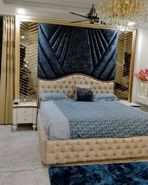 A master bedroom designed for pure luxury and comfort. The deep blue velvet headboard creates a dramatic focal point, complemented by a stunning crystal chandelier and custom mirrored panels, proving that a bedroom can be both a restful sanctuary and a statement of style.