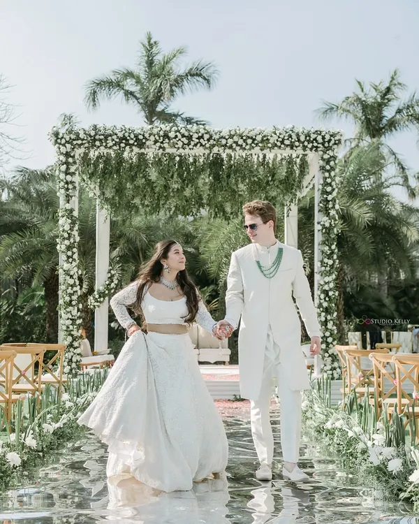A fairytale wedding in a pristine all-white theme. This shot captures the couple walking down a mirrored aisle, surrounded by lush greenery and palm trees.