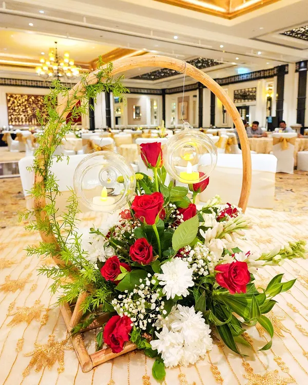 A unique and rustic centerpiece featuring a wooden hoop entwined with greenery. The arrangement of red and white flowers is accented with hanging glass orbs containing small candles, adding a touch of warmth and charm.