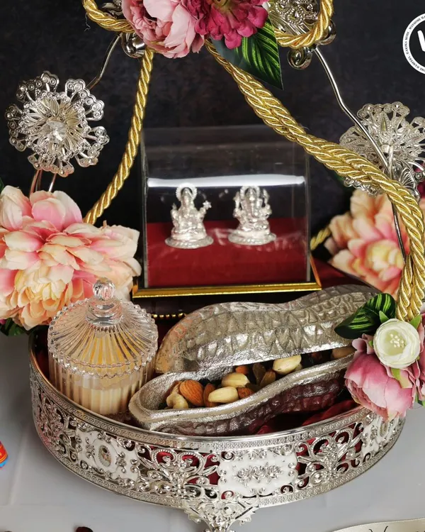 This opulent silver-plated tray is a centerpiece gift for Diwali. It features silver Lakshmi-Ganesh idols for blessings, a crystal jar for sweets, and a peanut-shaped container for dry fruits, all adorned with lush pink flowers.