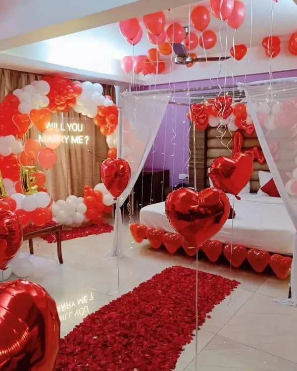 This is a complete proposal setup I designed for a client's room. It includes a 'Will you marry me?' neon sign, a red and white balloon arch, a rose petal walkway, and a decorated bed under a light-up canopy.