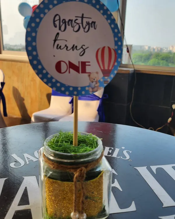 A charming table centerpiece for a hot air balloon themed first birthday. The personalized "Agastya turns ONE" sign is placed in a jar filled with glitter and moss, adding a touch of rustic charm.