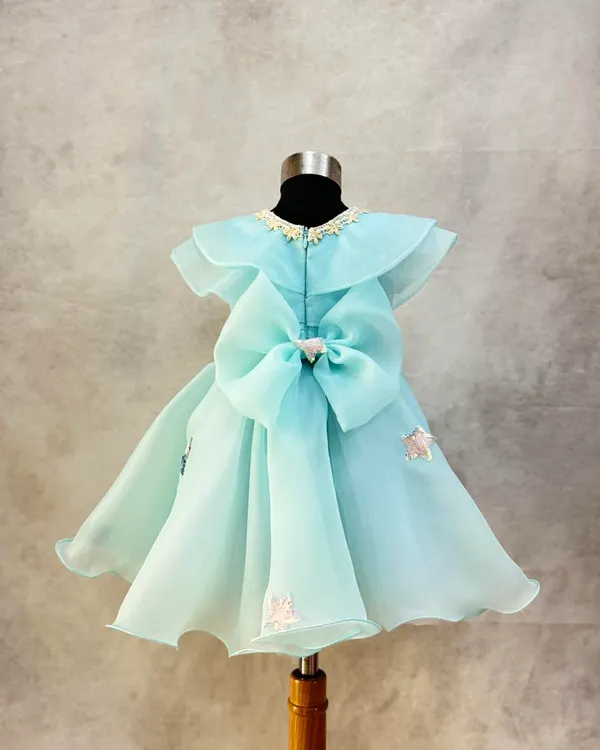 A custom dress in a beautiful shade of aqua organza. The design features a Peter Pan collar, delicate floral embroidery, and a large, statement bow at the back.