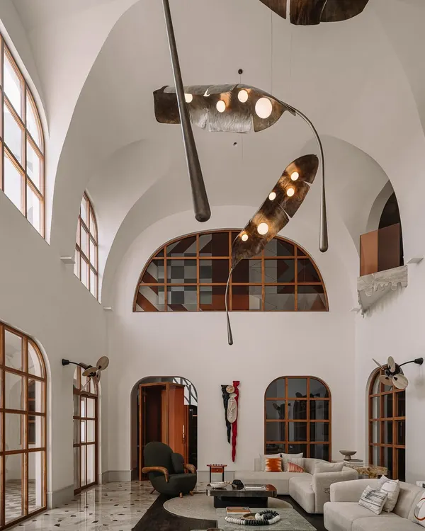 Sunlight streams into the double-height living room at Rehethaan, highlighting the grand scale, arched windows, and the massive, sculptural frond-like light installation. This space was designed to feel open, airy, and connected to the outdoors.