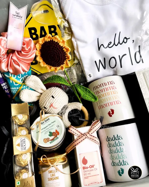 A perfect hamper for the parents-to-be, featuring 'Momma' and 'Dadda' mugs, a 'Hello World' onesie, skincare oil for the mom, and a cute crochet rattle for the baby.