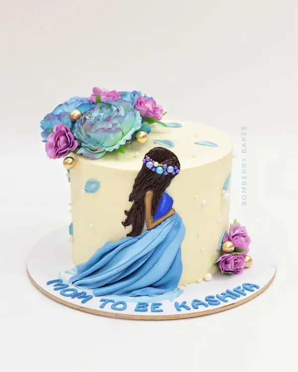 A beautiful "Mom to Be" cake featuring a hand-painted figurine of an expecting mother in a flowing blue dress, surrounded by delicate flowers.