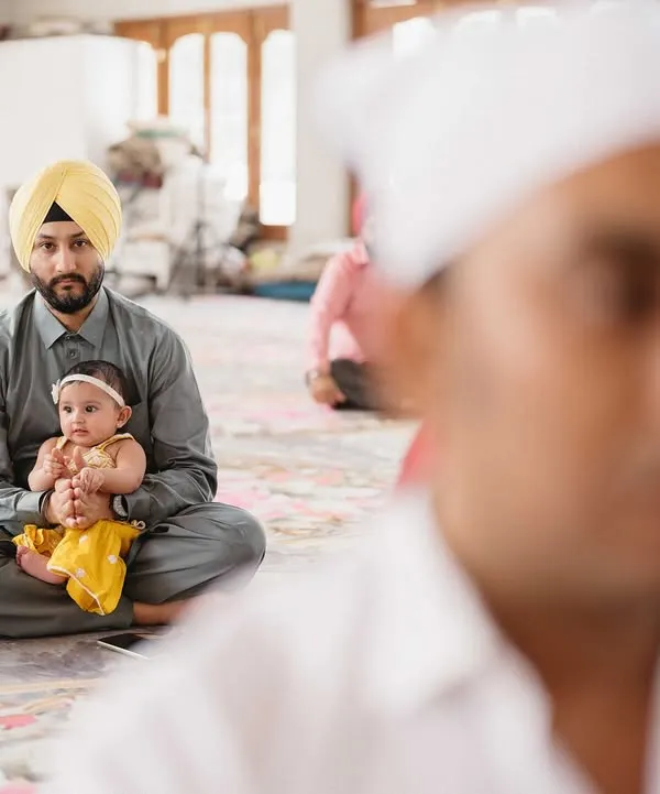 A father holds his young child during the Anand Karaj ceremony. This photo, shot with a shallow depth of field, captures a tender, multi-generational moment of faith and family.