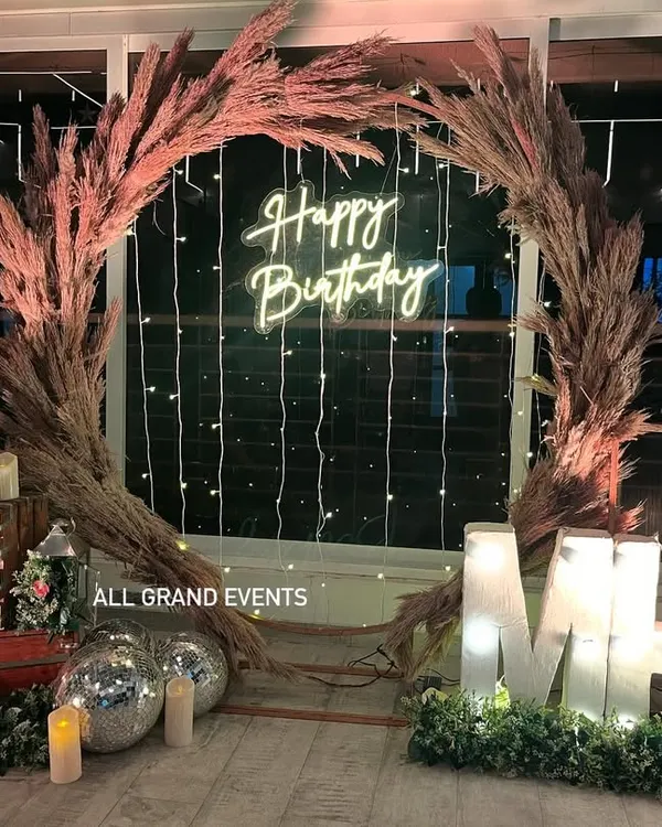 A beautiful rustic birthday backdrop featuring a large wreath made of pampas grass. The setup is personalized with light-up marquee initials and complemented by a neon sign and fairy lights.