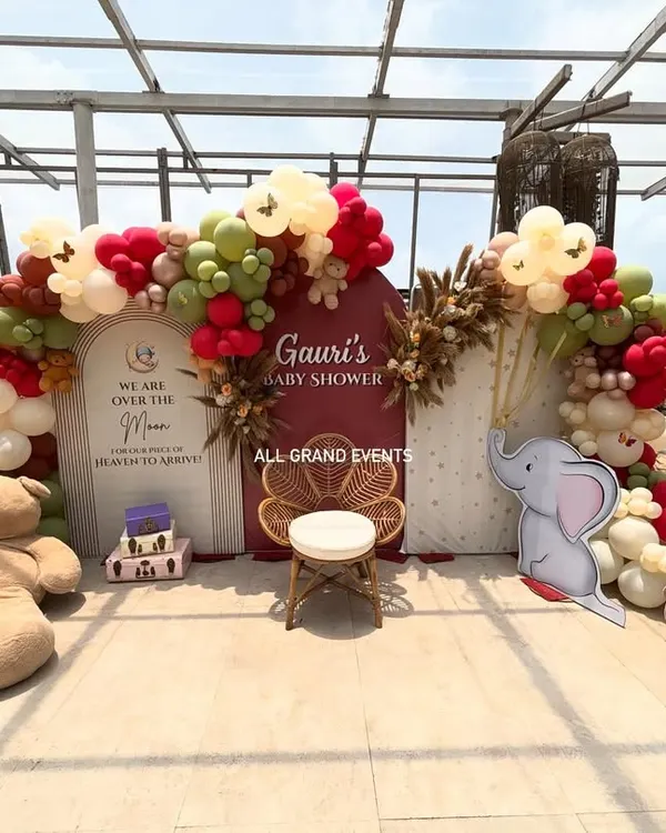 A beautiful 'Over the Moon' themed baby shower for Gauri. The setup includes two arched backdrops, a custom balloon garland in earthy red and green tones, and cute teddy bear and elephant props.