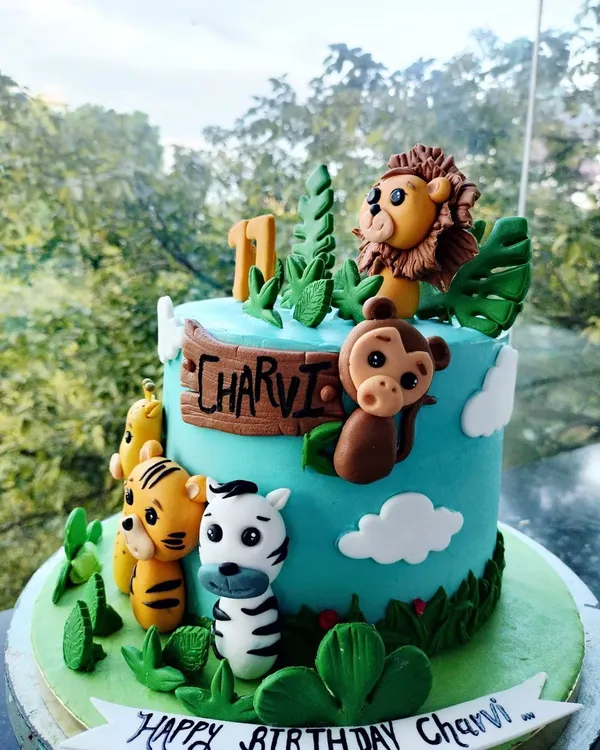 A vibrant jungle-themed birthday cake for Charvi's first birthday. This creation features adorable, handcrafted fondant animals like a lion, tiger, zebra, and monkey, all set against a bright blue sky and lush green leaves.