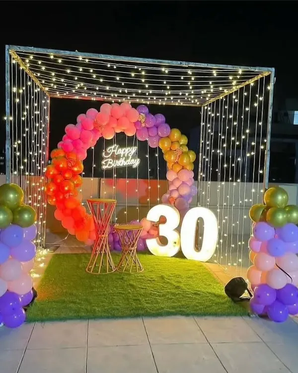 A vibrant 30th birthday setup on a terrace. This features a canopy of fairy lights, a colorful circular balloon garland, a 'Happy Birthday' neon sign, and large light-up numbers, all set on an artificial grass carpet.