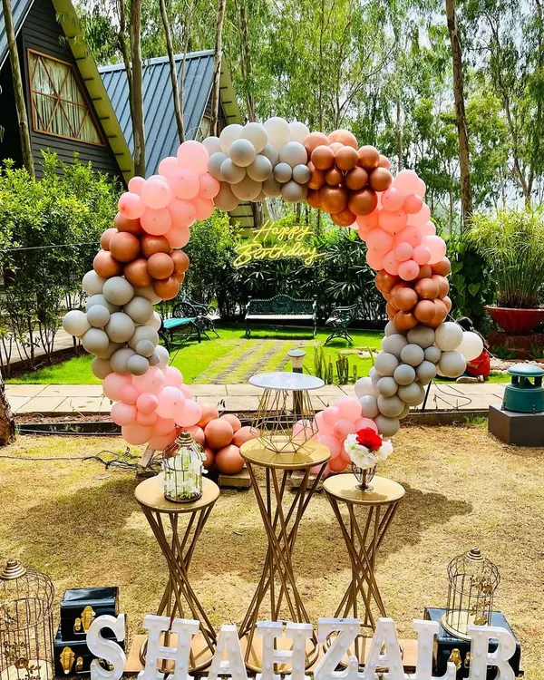 A beautiful daytime garden party featuring a circular balloon arch in muted tones of pink, grey, and bronze. The setup includes elegant dessert stands and personalized name lettering.