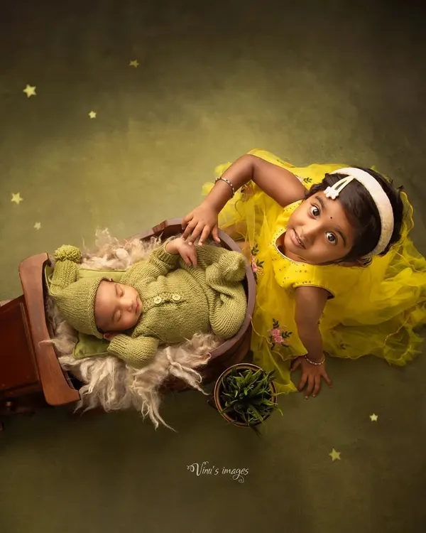 Big sister proudly watches over her sleeping newborn sibling. This creative shot uses a miniature wooden cart as a prop, adding a whimsical touch to this sibling and newborn portrait.