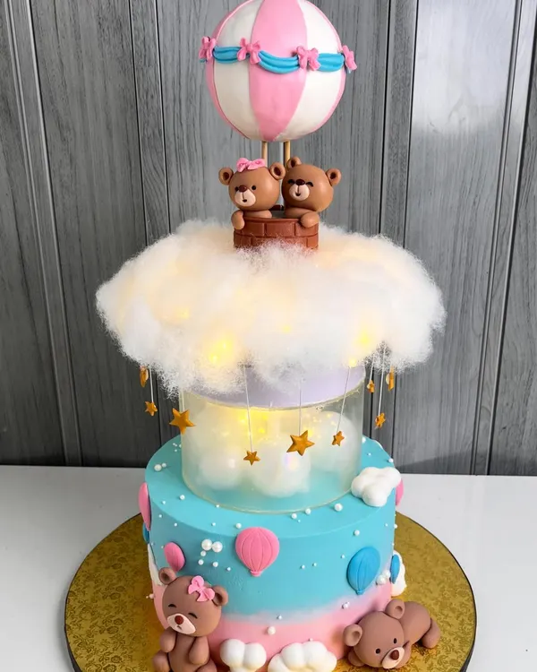 A magical two-tiered baby shower cake featuring teddy bears in a hot air balloon. The middle tier is illuminated with fairy lights behind a cloud of cotton, creating a dreamy, glowing effect.