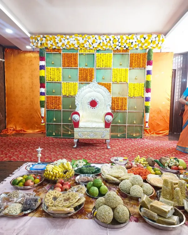 A vibrant Shreemantha setup featuring a checkered backdrop of fresh marigold flowers and an ornate silver chair for the mother-to-be, with traditional offerings displayed in front.