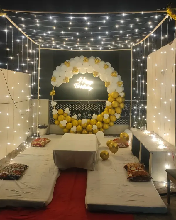 A beautiful cabana-style terrace decoration for a birthday, featuring a canopy of fairy lights, low seating with mattresses, and a crescent-shaped balloon arch.