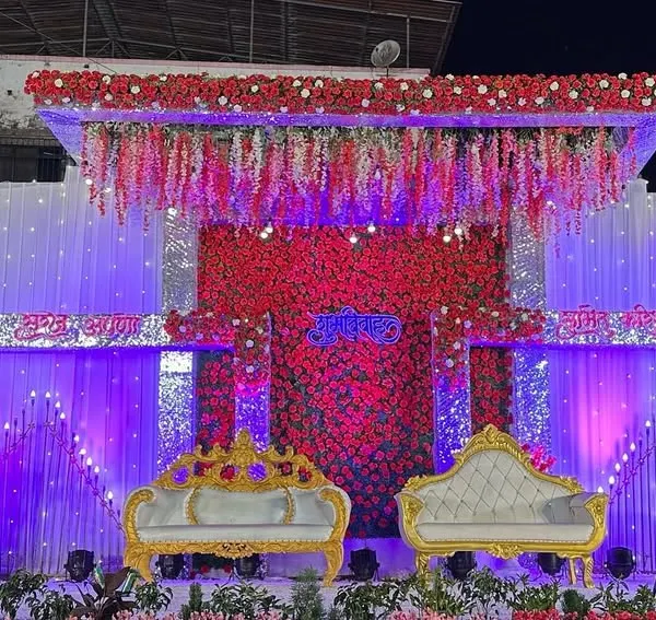A grand reception stage featuring a massive red and white floral backdrop. The design incorporates hanging floral strings and ornate, royal-style seating to create a look of traditional grandeur.