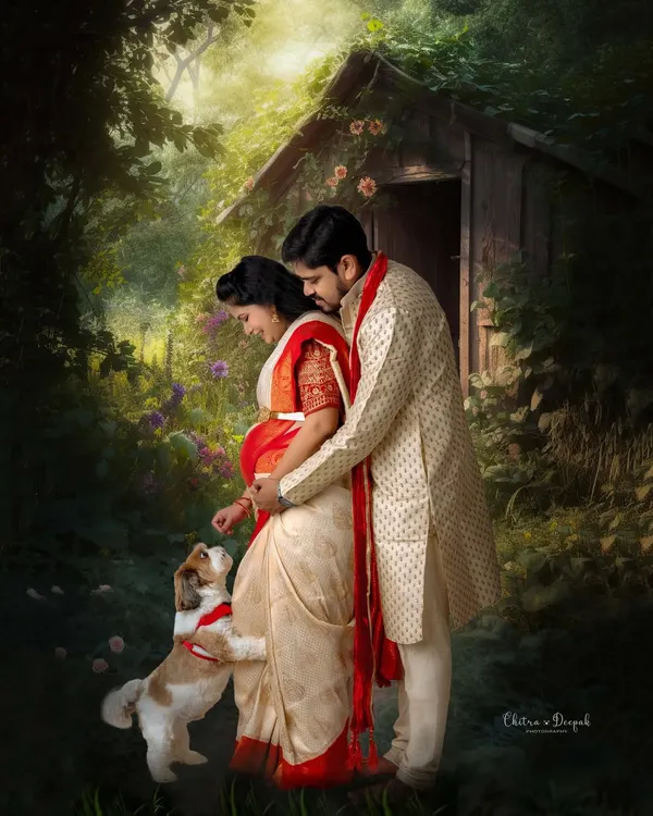 An artistic and romantic maternity portrait of a couple in traditional Indian attire, sharing a tender moment with their beloved pet in a magical, painterly forest setting.