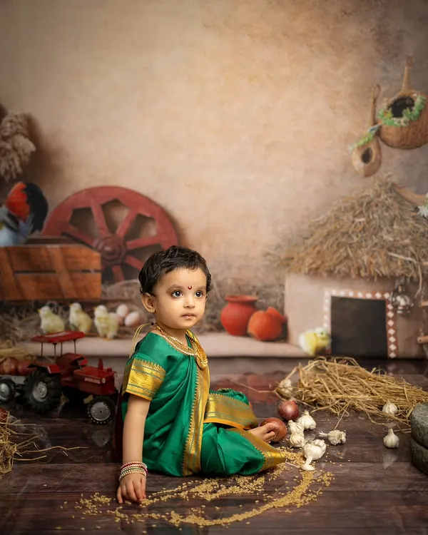 A thoughtful moment captured in our rustic Maharashtrian village set. The details, from the hay hut to the toy tractor, create an authentic backdrop for this traditional child photoshoot.
