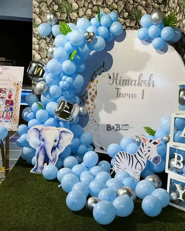 A cheerful and bright first birthday setup with a blue animal theme. This decoration includes a large balloon arrangement, cutouts of a baby elephant and zebra, and matching "BABY" display blocks.