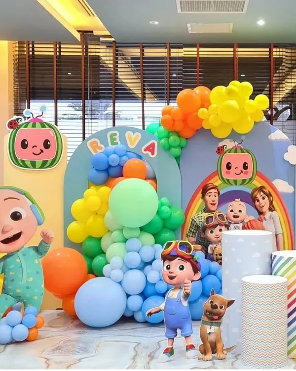 A bright and cheerful Cocomelon themed birthday party for Reva. This wide shot shows the full setup, including multiple character cutouts, themed plinths, and a rainbow of colorful balloons.