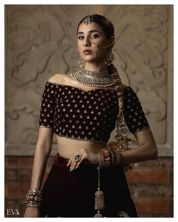 A powerful and elegant bridal fashion shot, showcasing a model in a deep maroon velvet lehenga. The off-shoulder blouse, intricate jewellery, and confident pose against a textured backdrop highlight our expertise in creating high-fashion ethnic lookbooks.