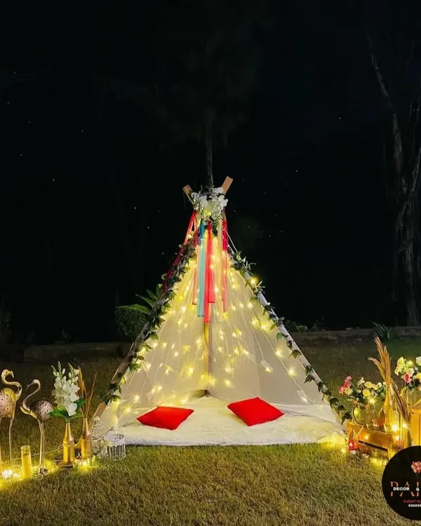 A magical teepee tent setup in a garden at night. It's decorated with fairy lights, flowers, and ribbons, creating a perfect spot for a romantic date.