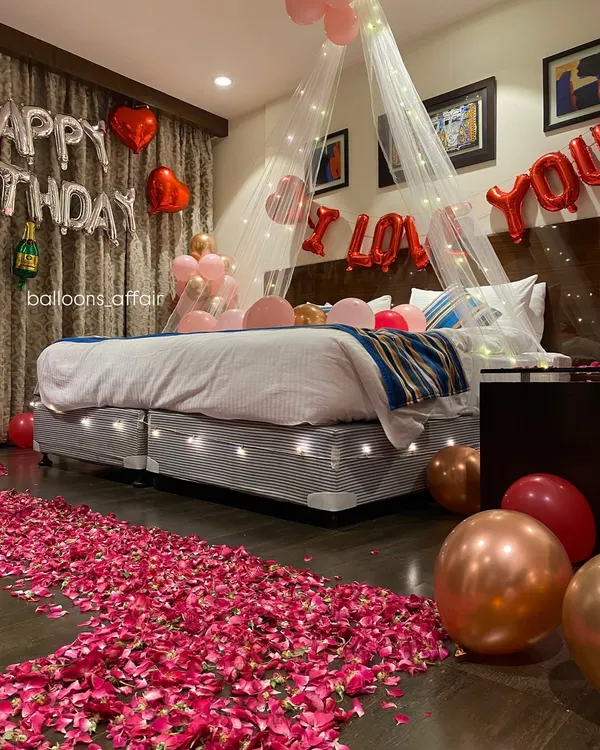 I designed this hotel room for a truly romantic birthday surprise. It features a beautiful rose petal pathway leading to a bed adorned with a delicate canopy and fairy lights, complemented by rose gold and red balloons.